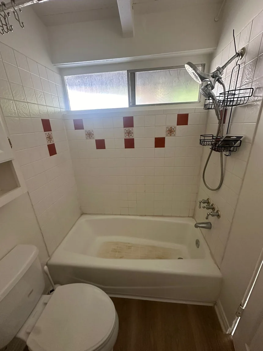 A white tiled bathroom with a band of red and patterned decorative tiles, and a bathtub stained brown at the base.