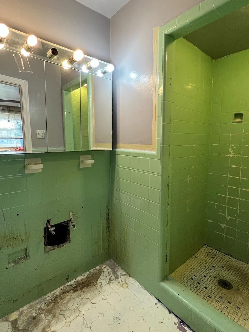 A 1950s bathroom tiled in green to chest height, with a green shower stall, moldy grout and a stained hexagonal tile floor.