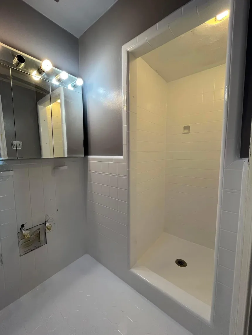 The same bathroom refinished in white: walls, shower stall and hexagonal floor, with the mirrored cabinet and light bar untouched.