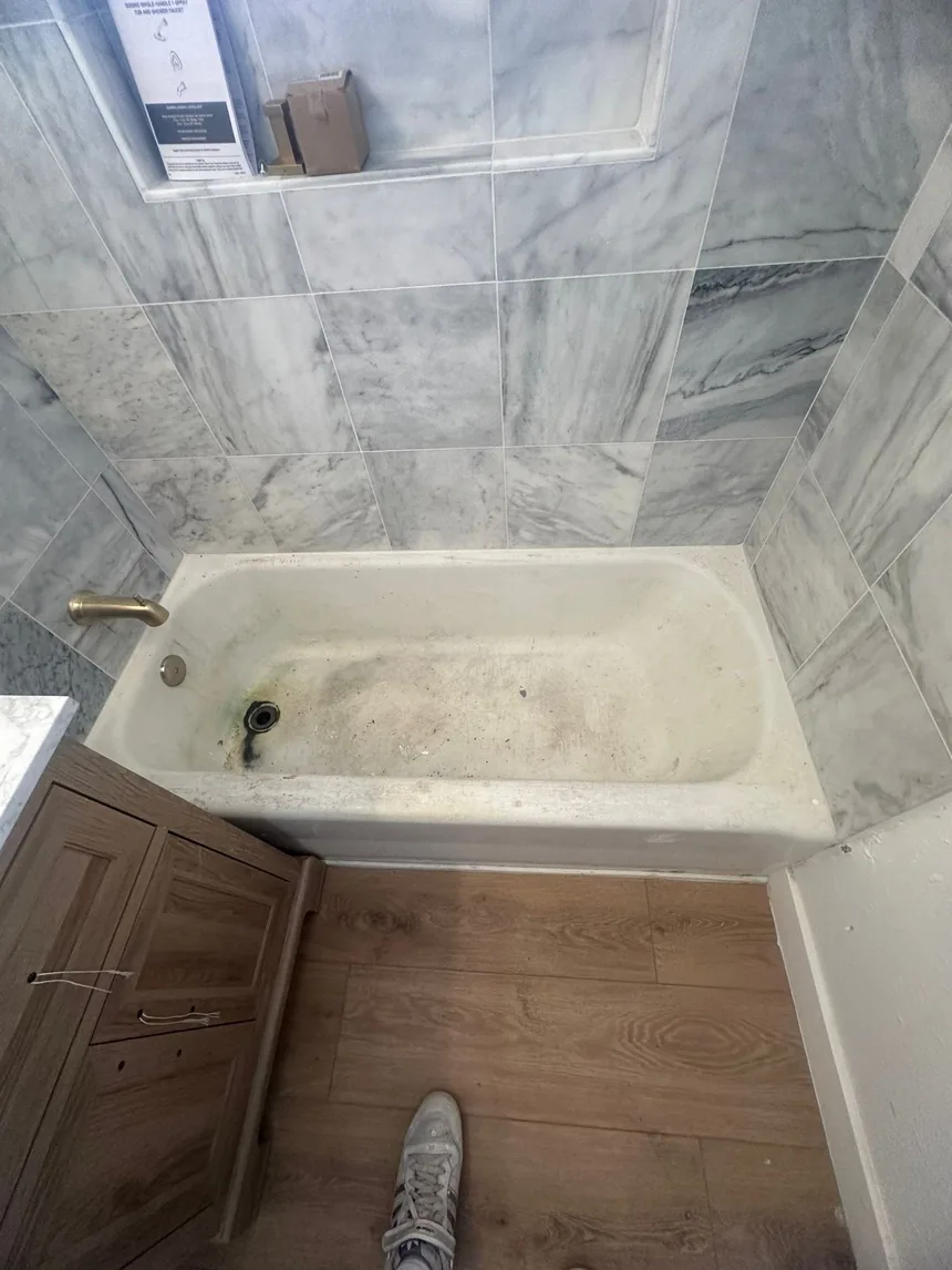 A bathtub with heavy gray staining across the base, in a bathroom tiled in large veined marble-effect tile.