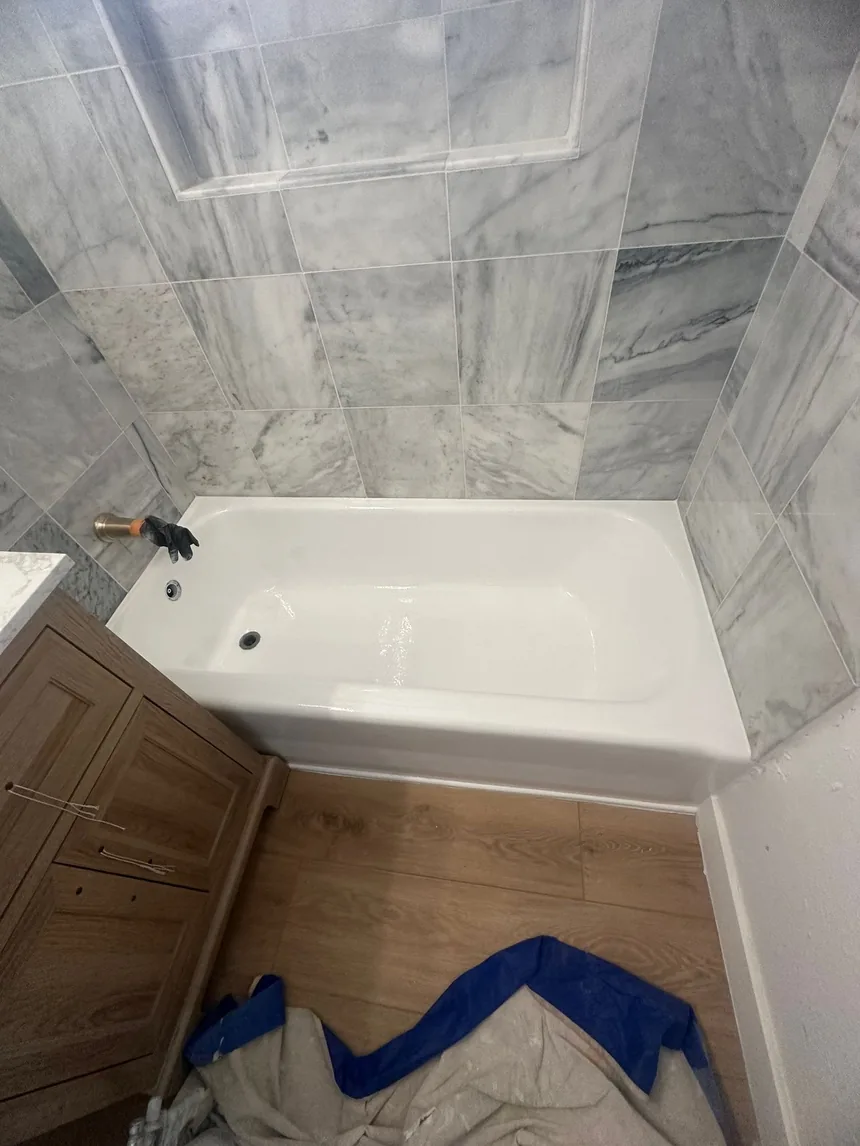 The same bathtub refinished in gloss white, against the same marble-effect tile and wood vanity.