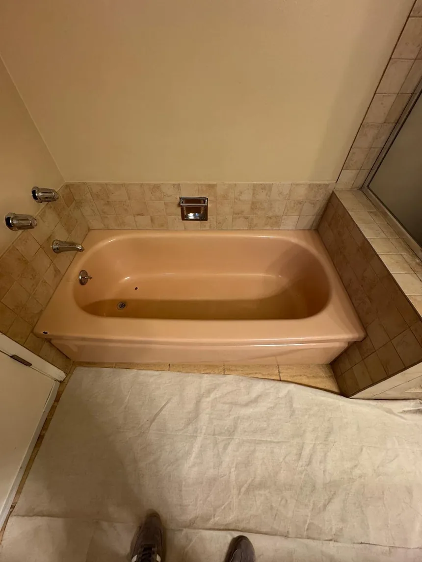 An original pink bathtub set into beige tile, with a dust sheet over the floor, before any work.
